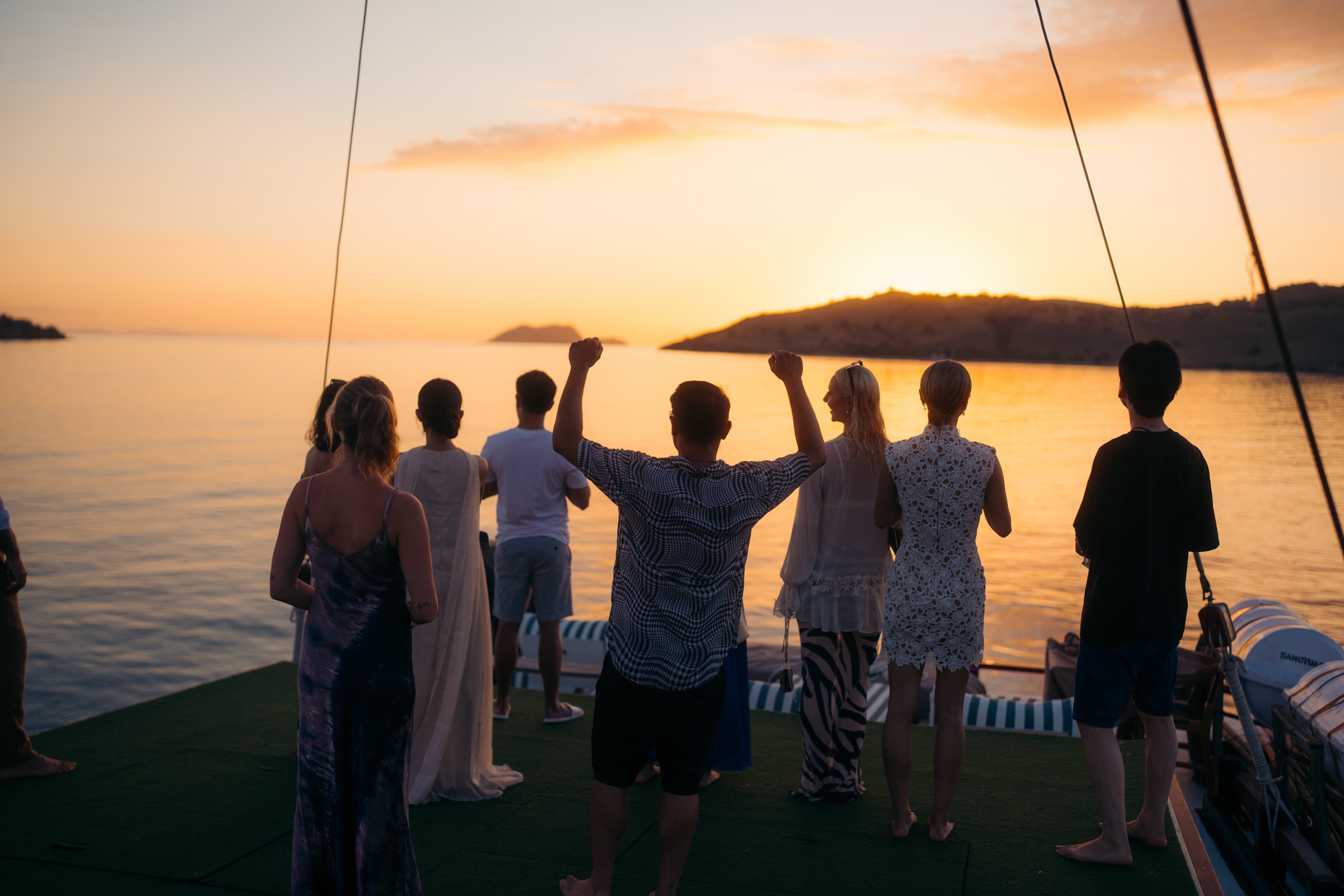 Guests on the deck of a phinisi, facing the sunset over the islands