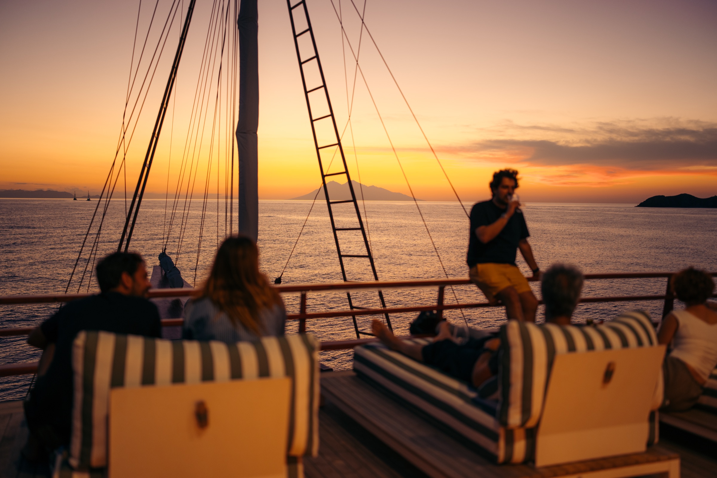 Guests on the deck of a phinisi at sunset, the rigging against an orange sky