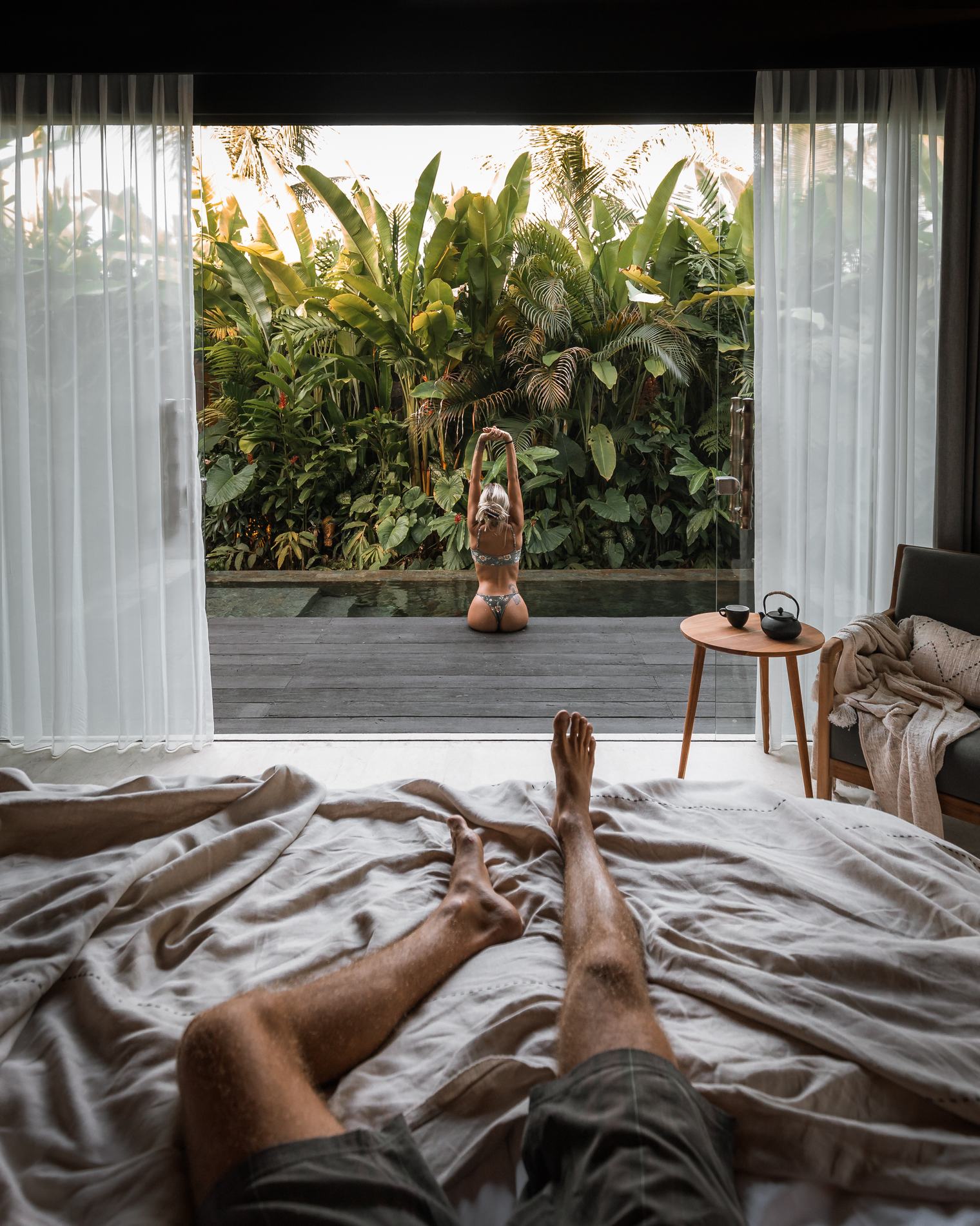 A view from the bed through open curtains to a woman stretching outside