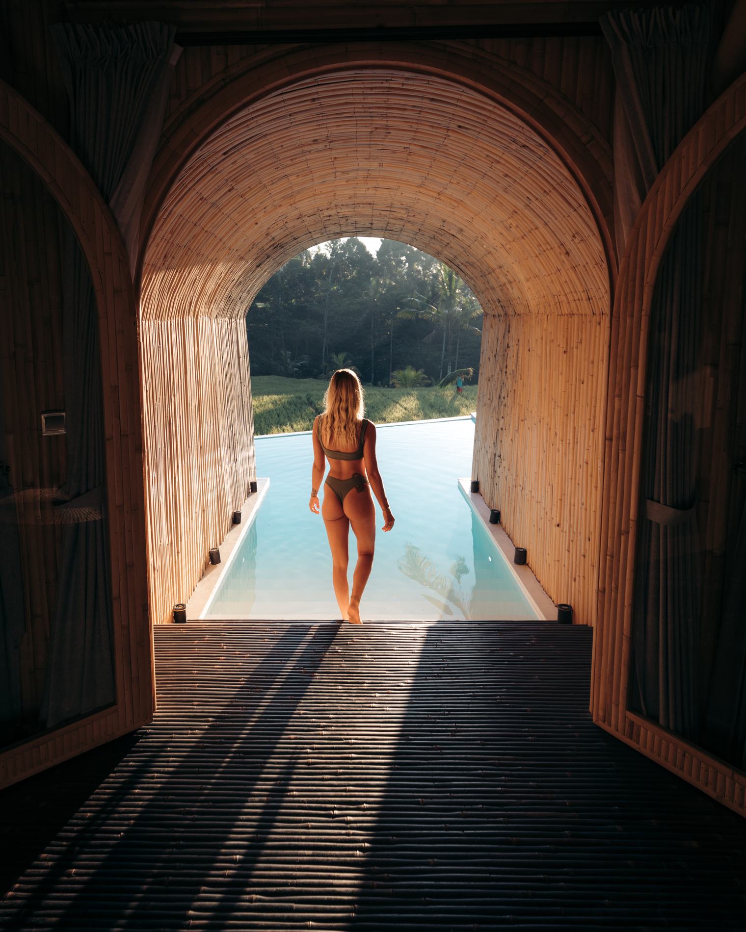 A woman walking through a bamboo arch toward a pool