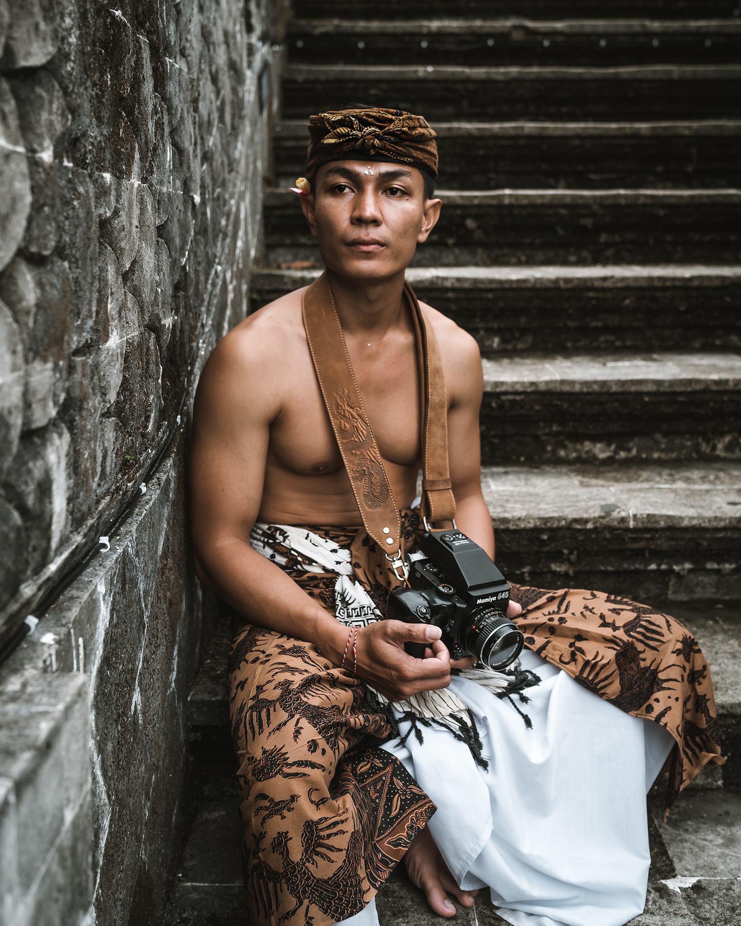 A man in a sarong sitting on temple steps with a camera on a strap