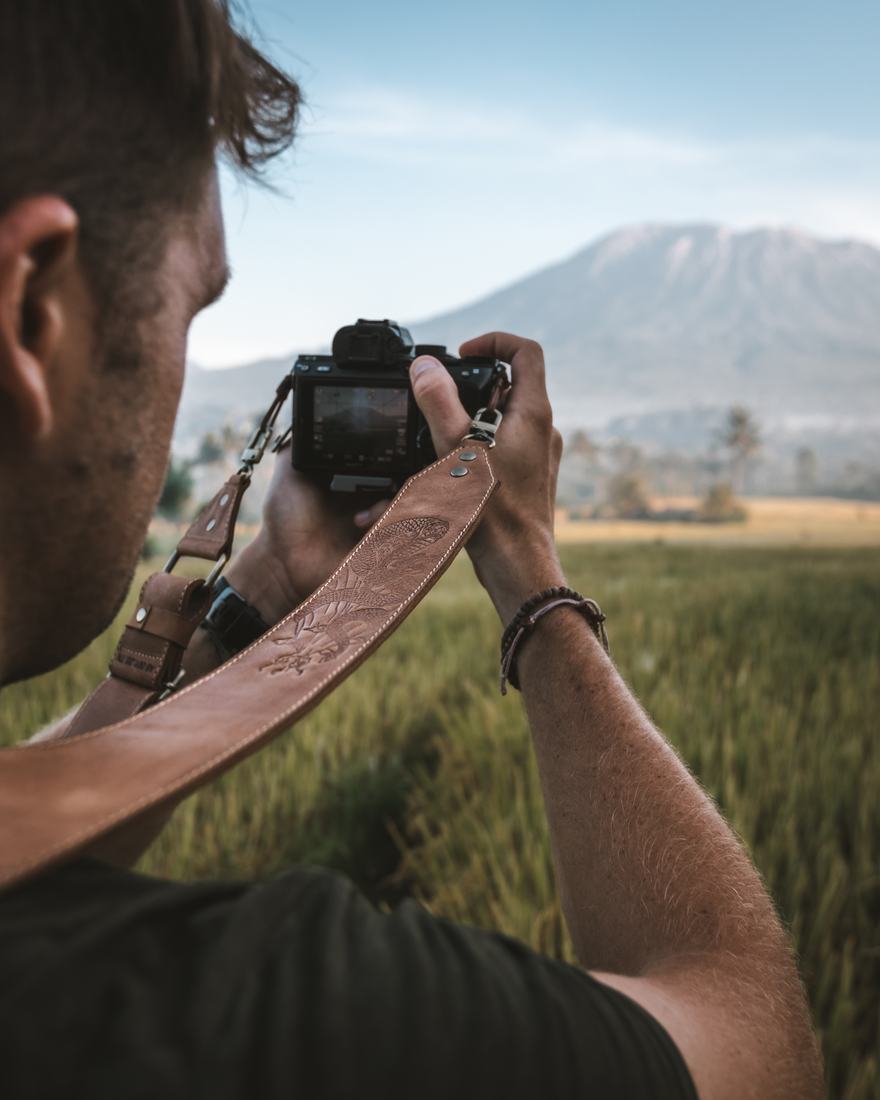 A photographer raising a camera on a leather strap in a rice field, a mountain behind