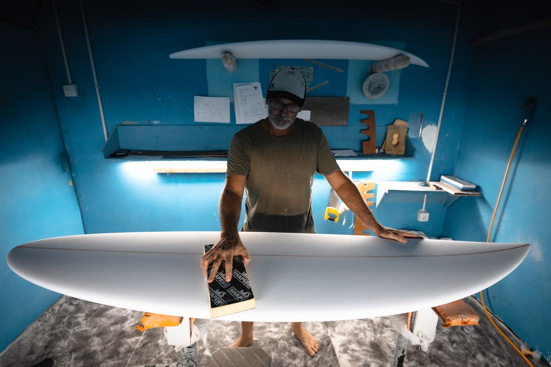 A shaper sanding a white surfboard in a blue-walled bay