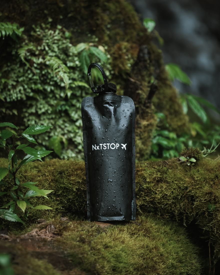A black Next Stop bottle standing on a mossy rock beside a waterfall