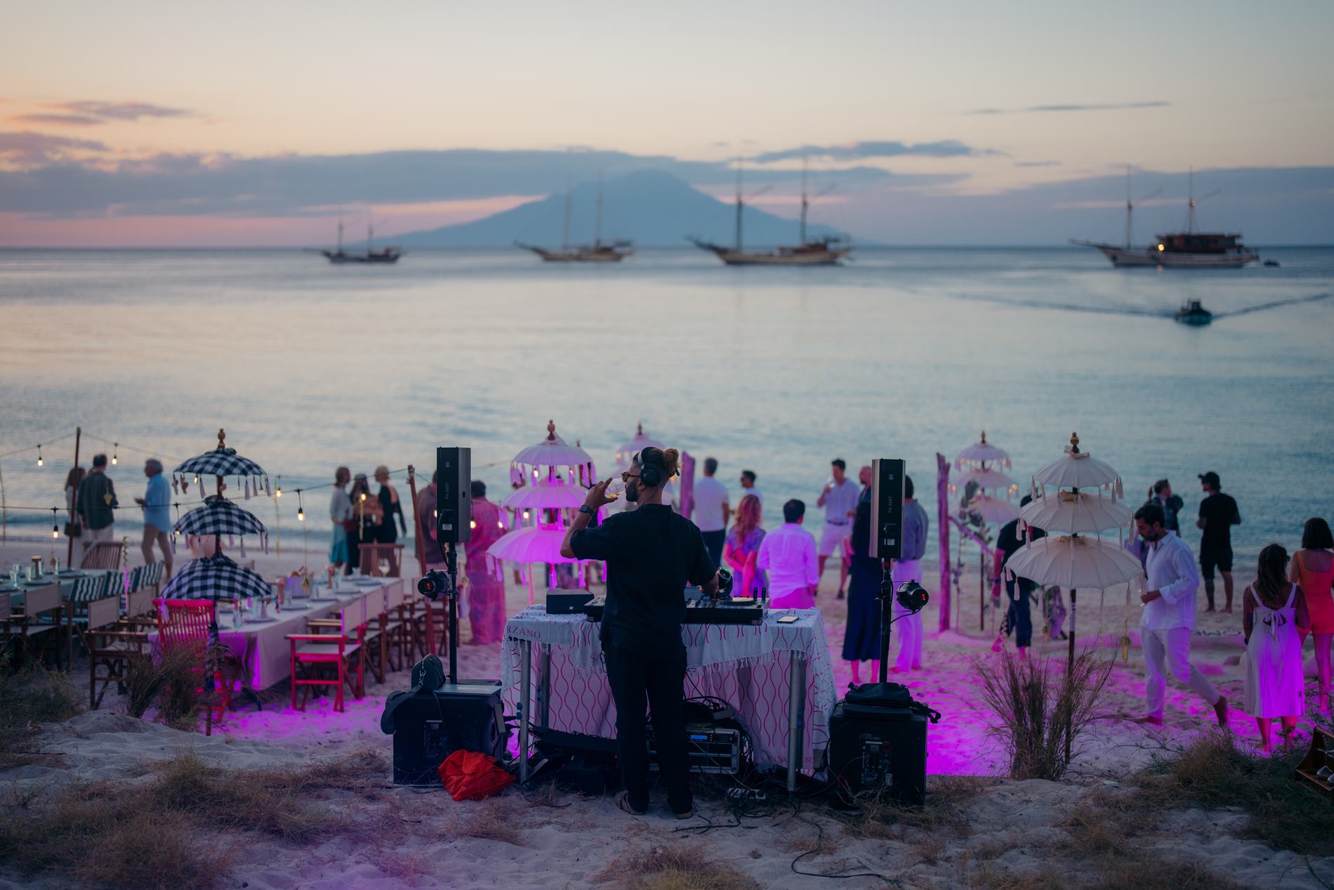 A beach party at dusk with a DJ, long tables and ships anchored offshore