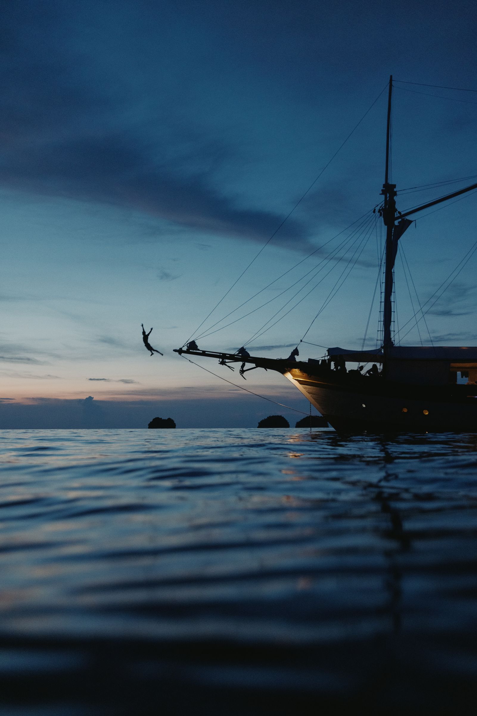 A wooden sailing ship at dusk under a deep blue sky, a figure swinging from its bowsprit