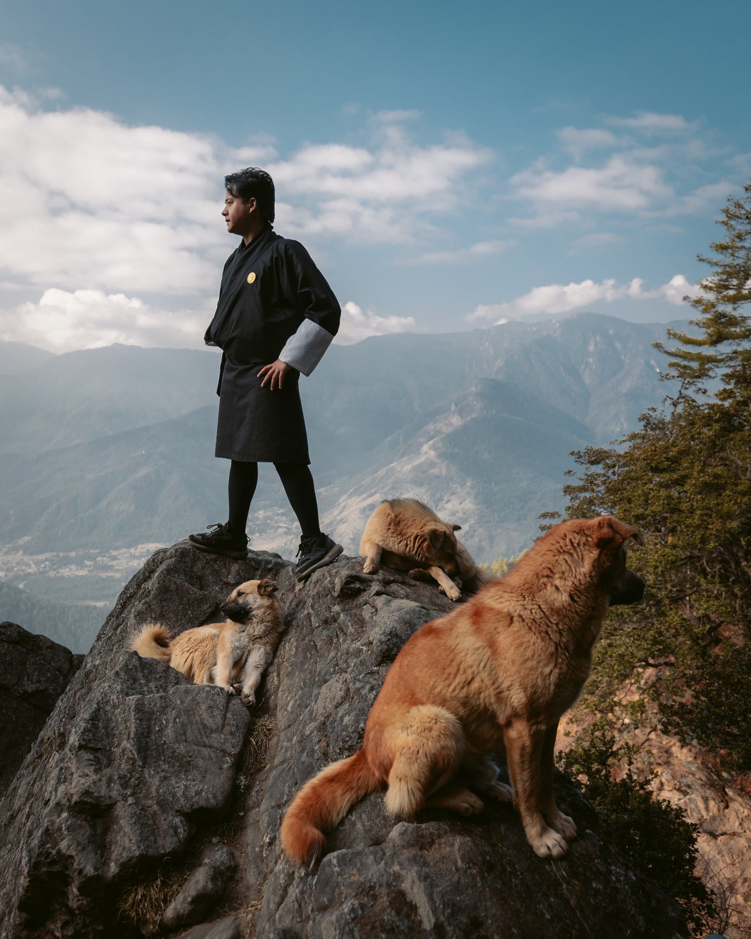 A man in traditional dress standing on a rock with two dogs, mountains behind