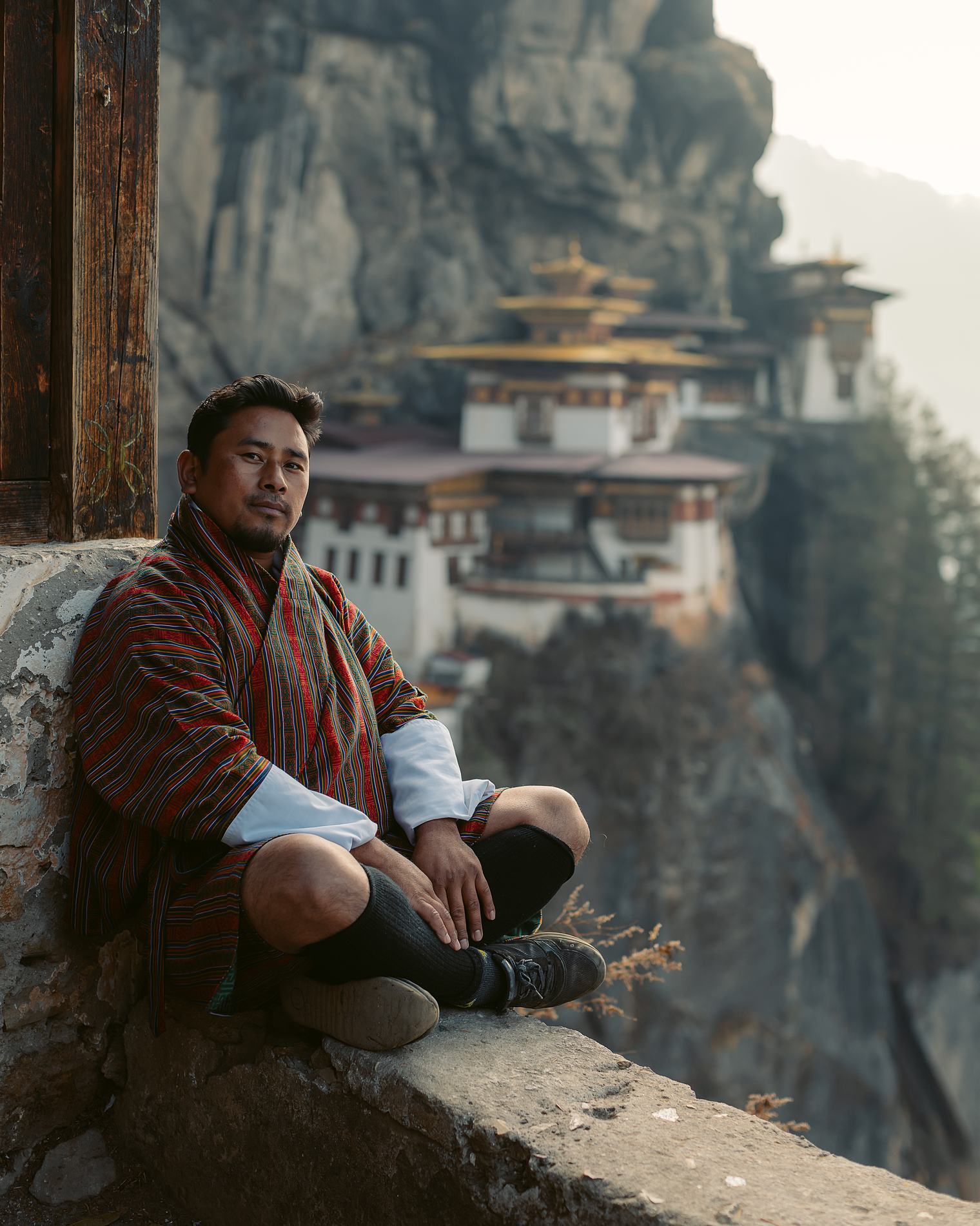 A man sitting against a stone wall, a cliffside monastery behind him