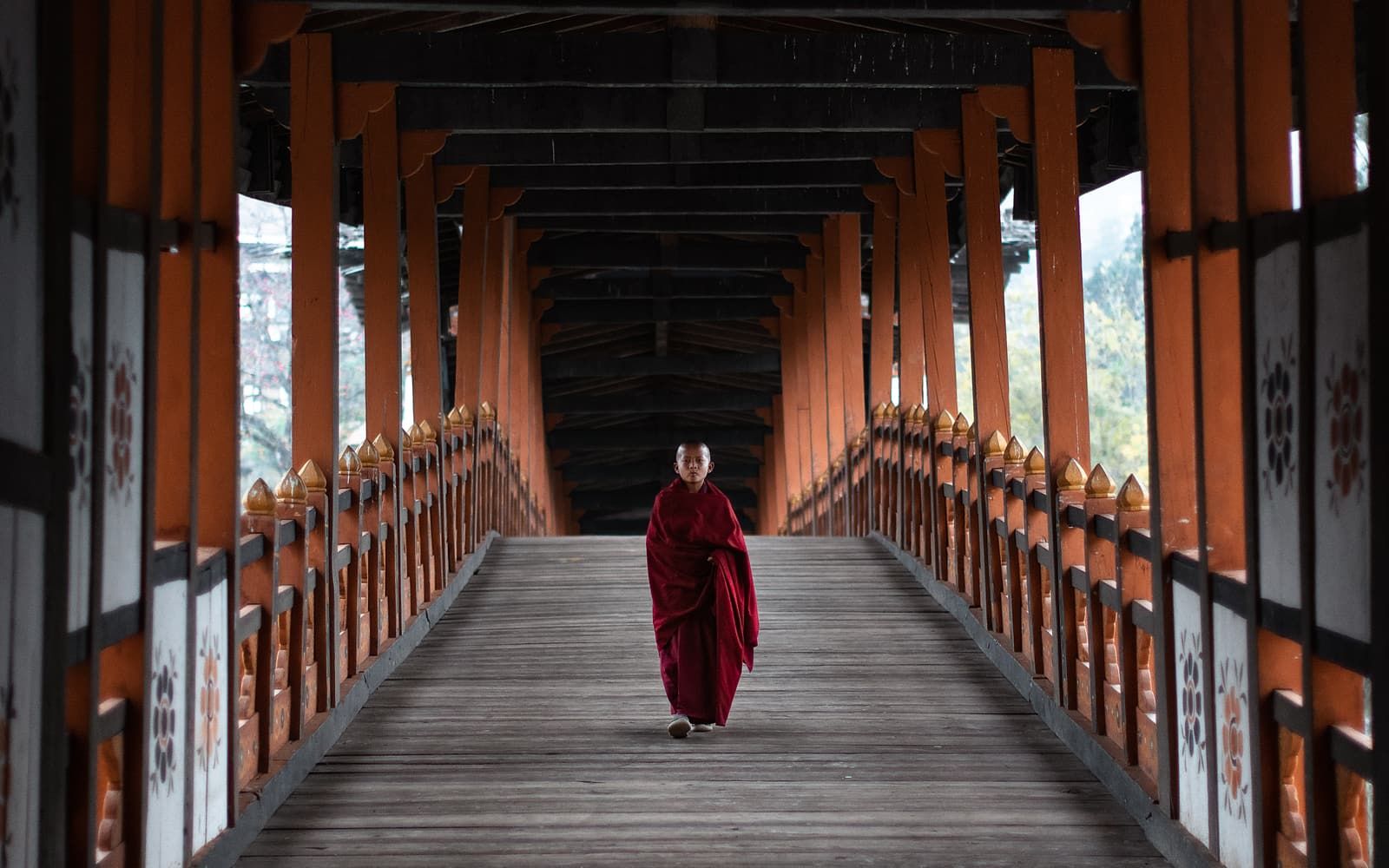 A monk in red robes walking along a covered wooden bridge