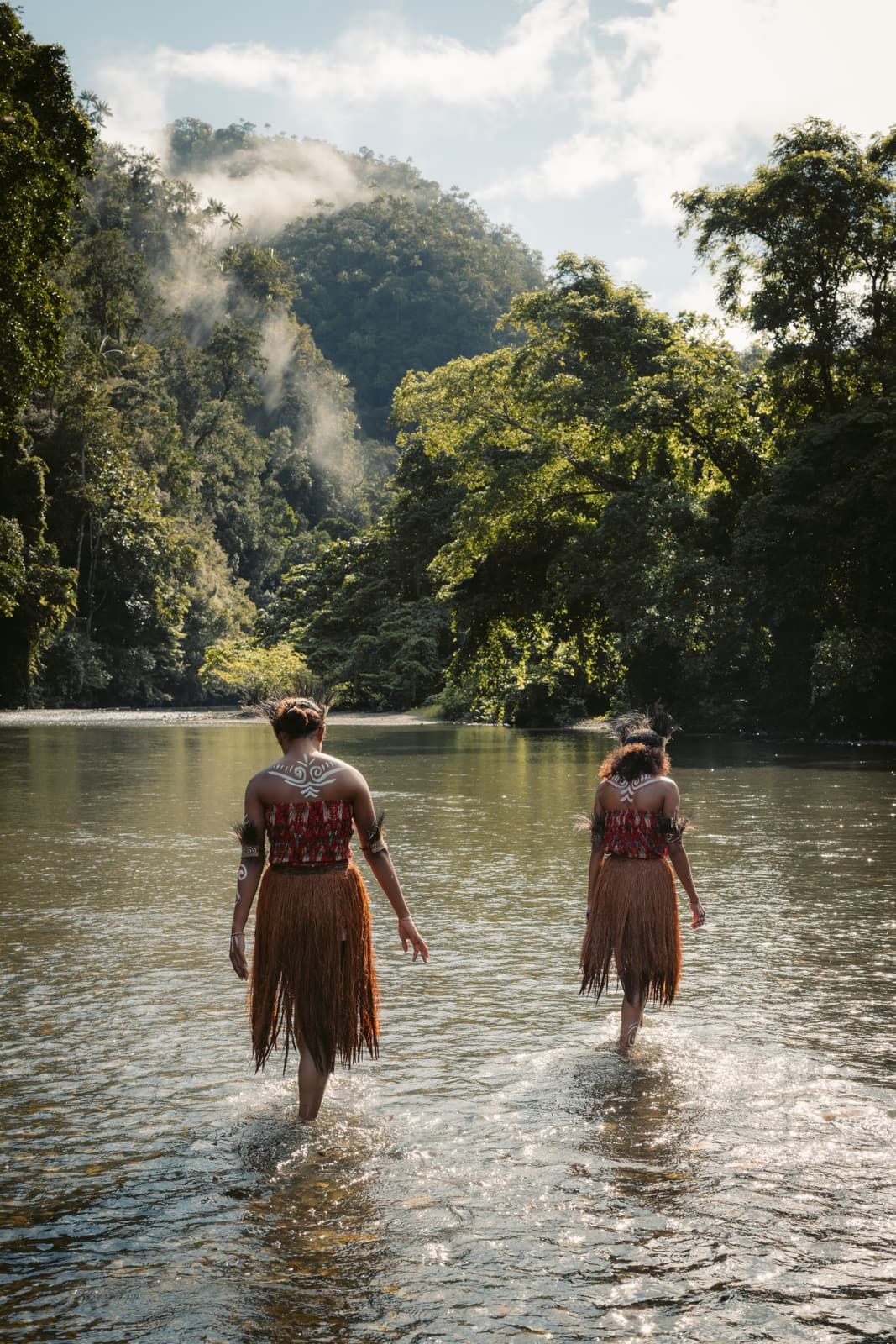 Two women in woven skirts wading up a shallow river toward misty forested hills