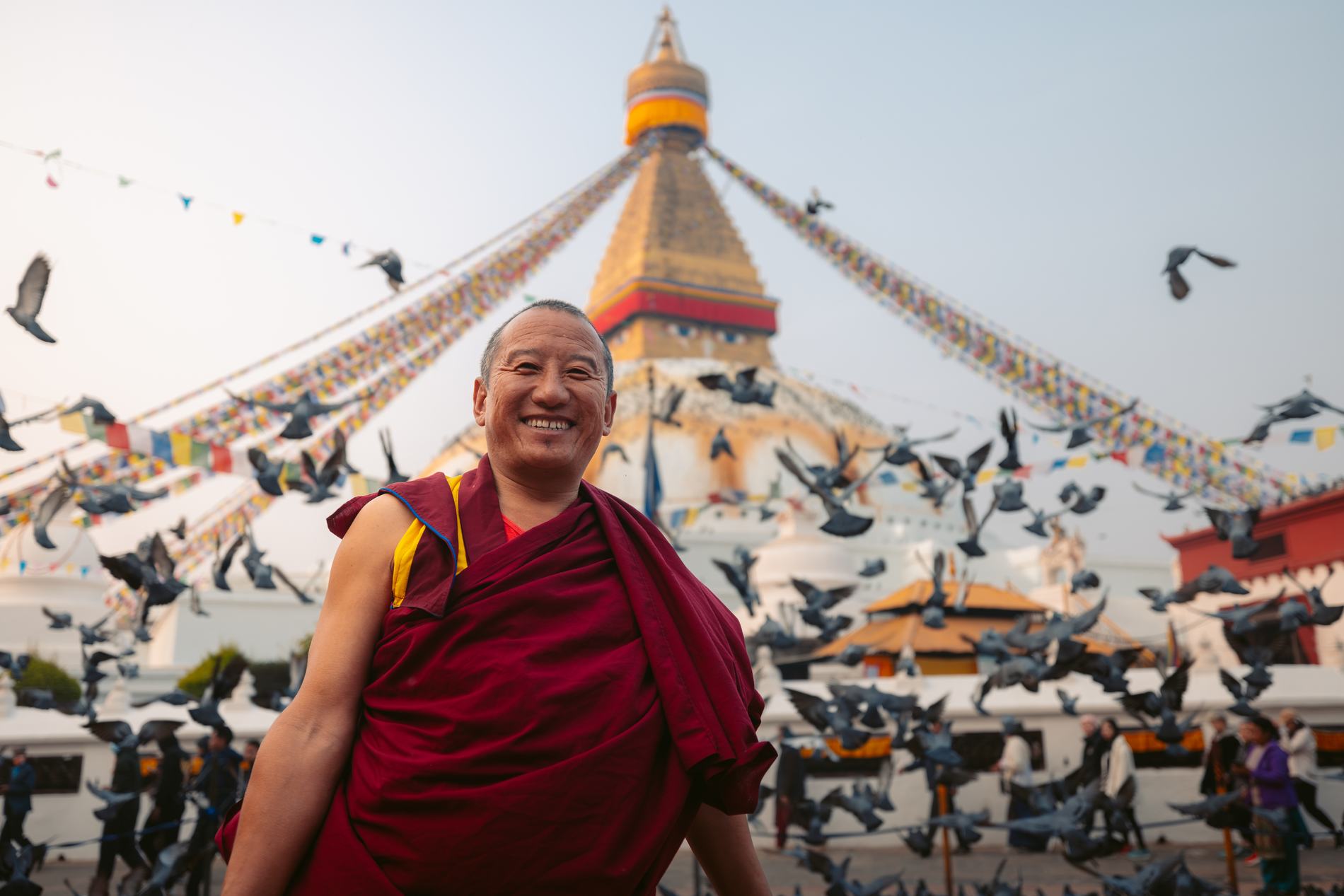 A smiling monk in maroon robes with pigeons and the stupa behind him