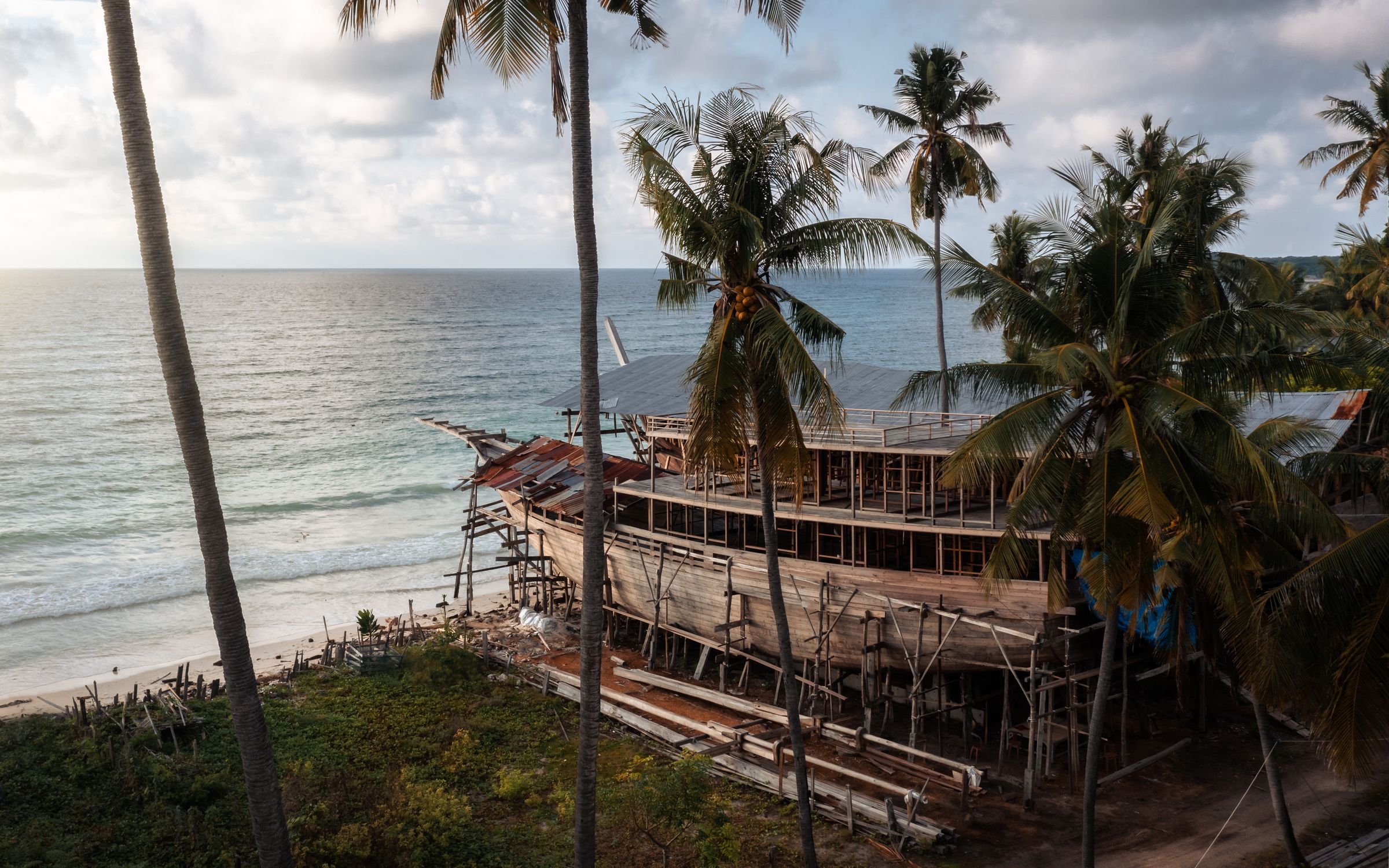 A wooden phinisi hull taking shape on a beach behind palm trees