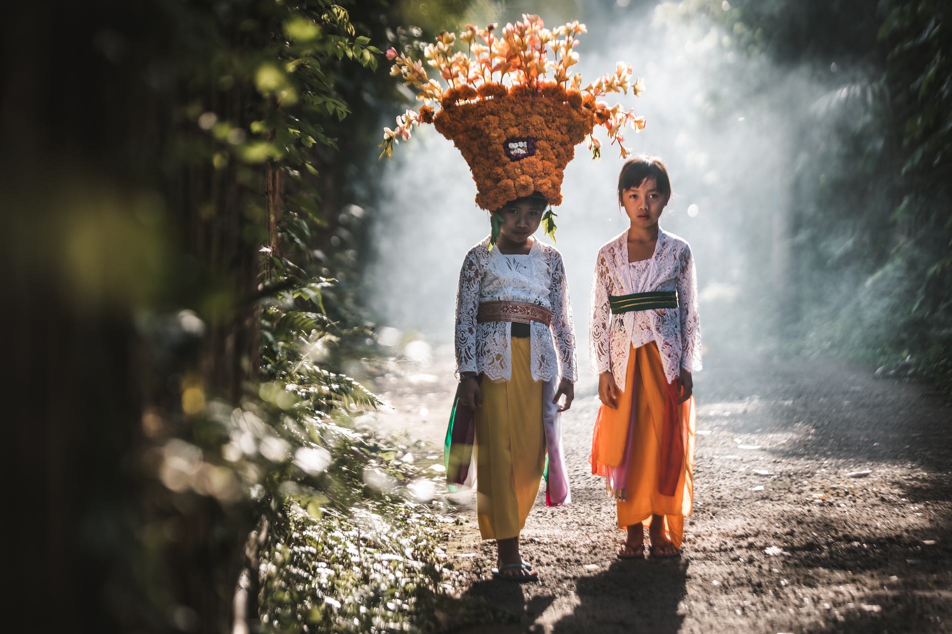 Two girls in traditional dress, one carrying an offering on her head